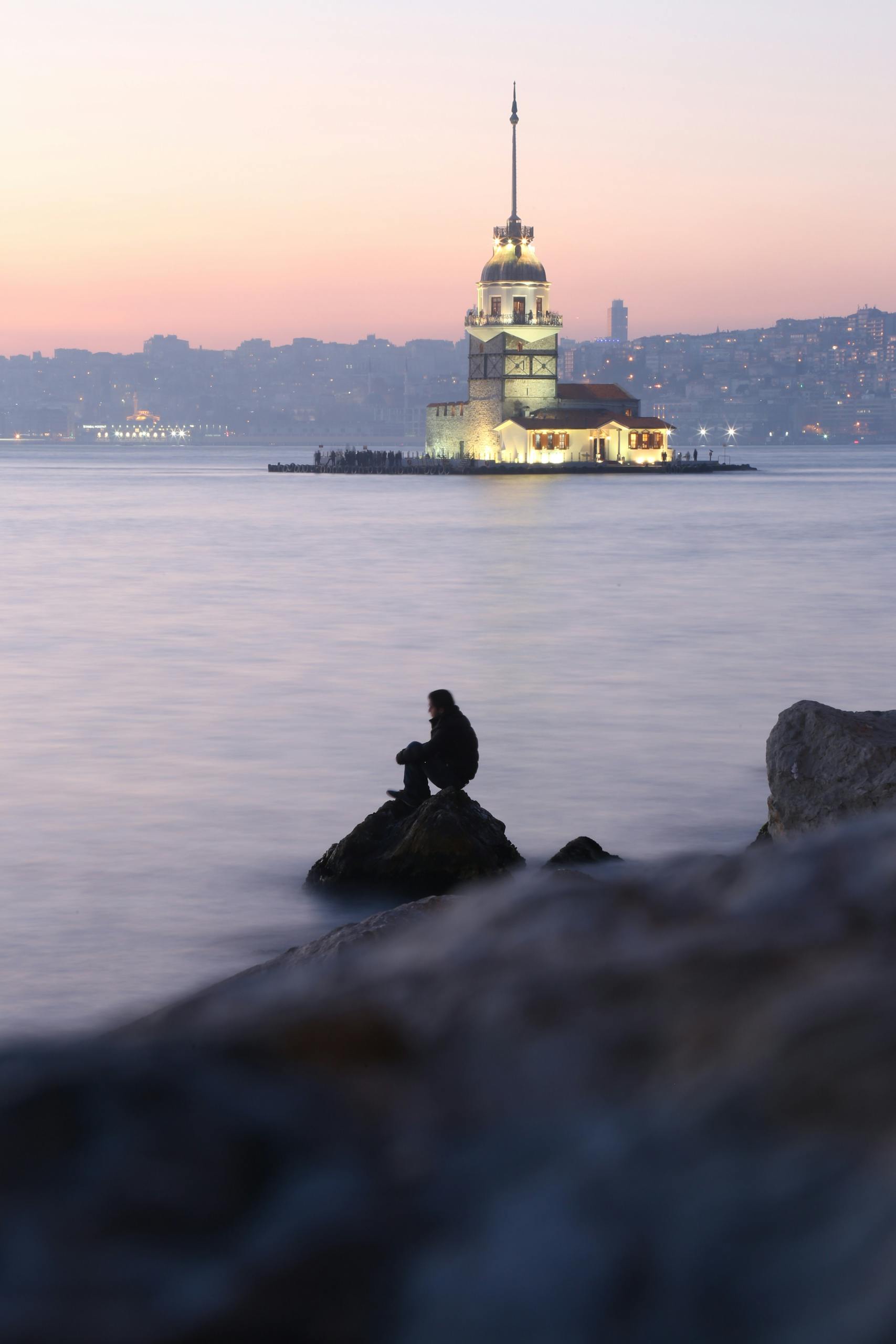 Silhouette of a person sitting by the sea with the illuminated Maiden's Tower in İstanbul at sunset.